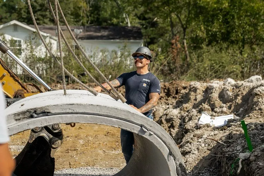 Construction worker in hard hat guiding a concrete pipe segment at a construction site.