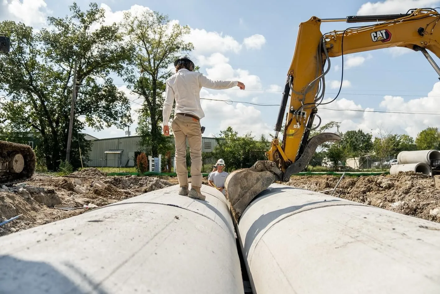 Man standing on large concrete pipes, pointing. Excavator in the background. Construction site.