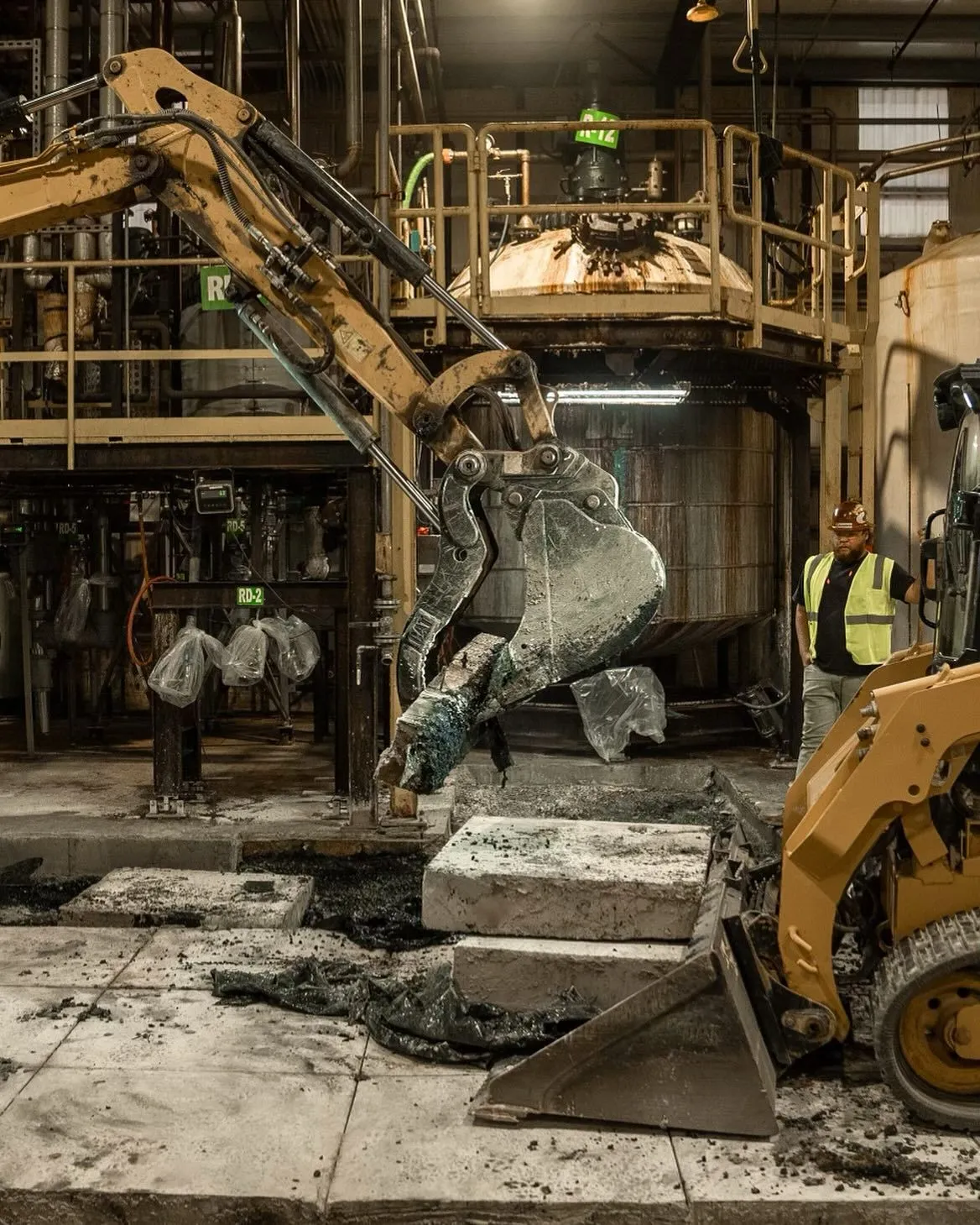 Yellow excavator scoops material on a factory floor. Worker in a reflective vest watches.