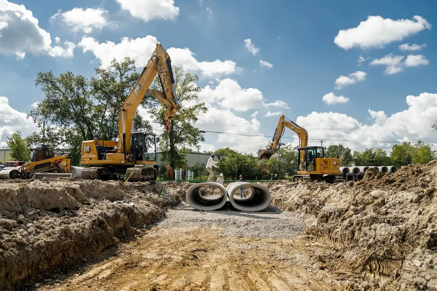 Construction site with excavators, large concrete pipes, and trenches under a blue sky.