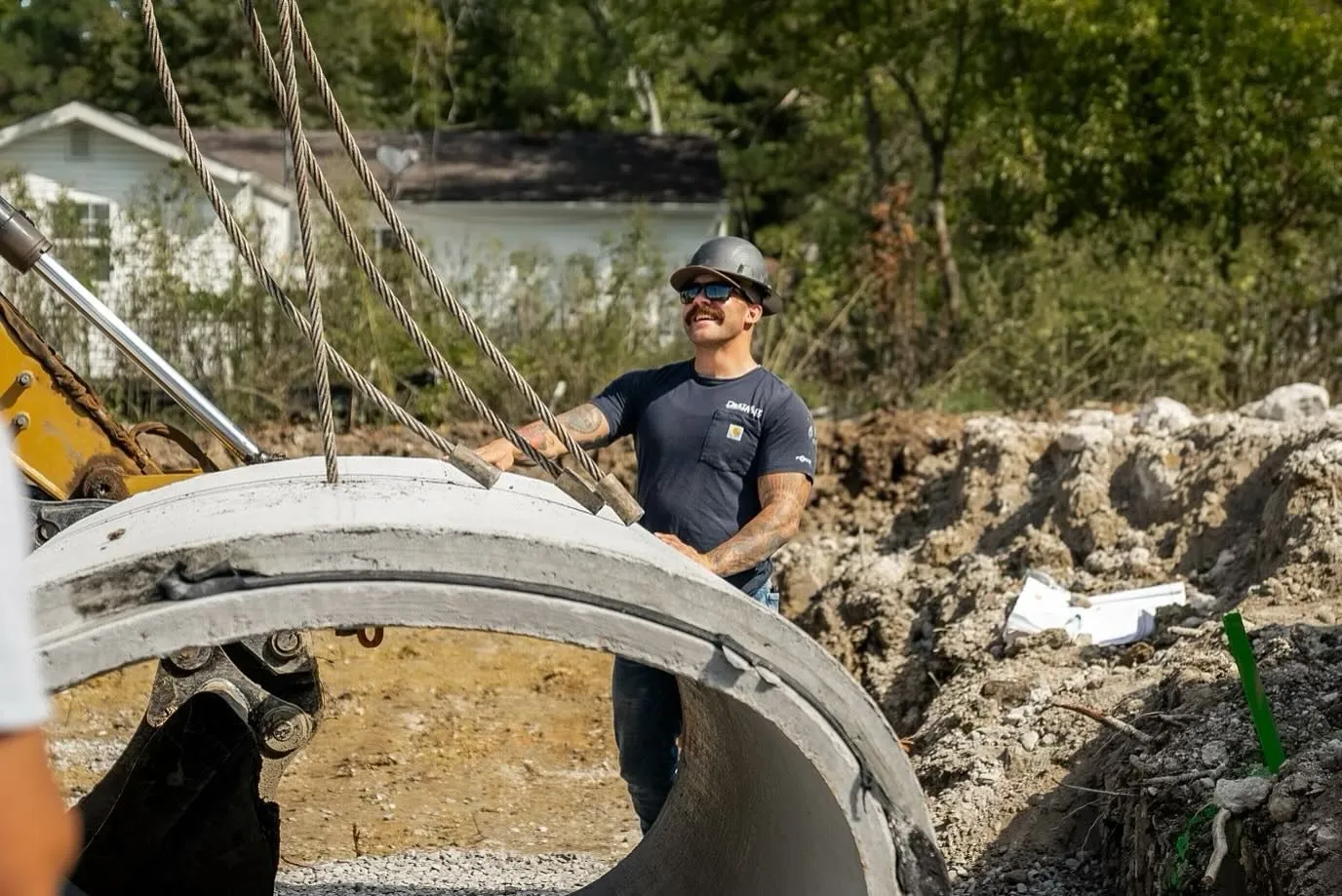 Construction worker guiding a concrete ring into a hole, using chains. Outdoor setting, daylight.