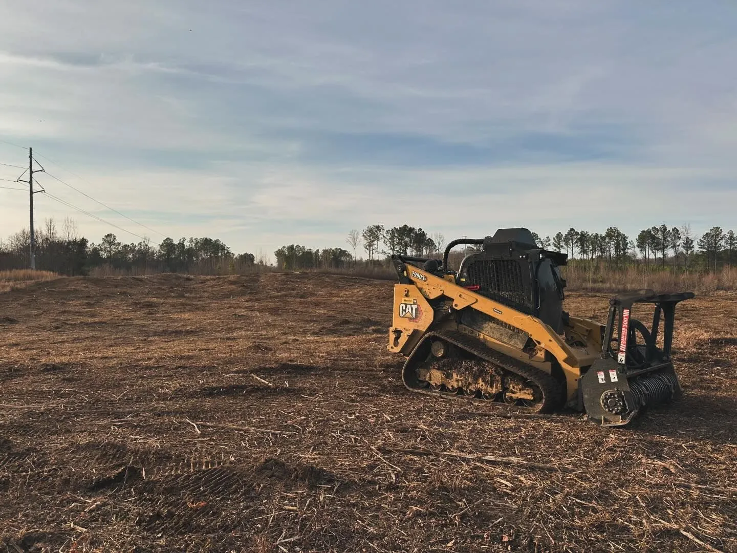 Yellow Caterpillar skid steer with brush cutter in a cleared field, cloudy sky.