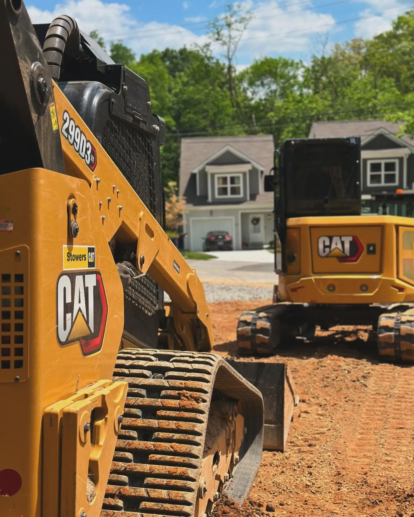 Yellow CAT construction vehicles on a dirt lot with houses in the background on a sunny day.