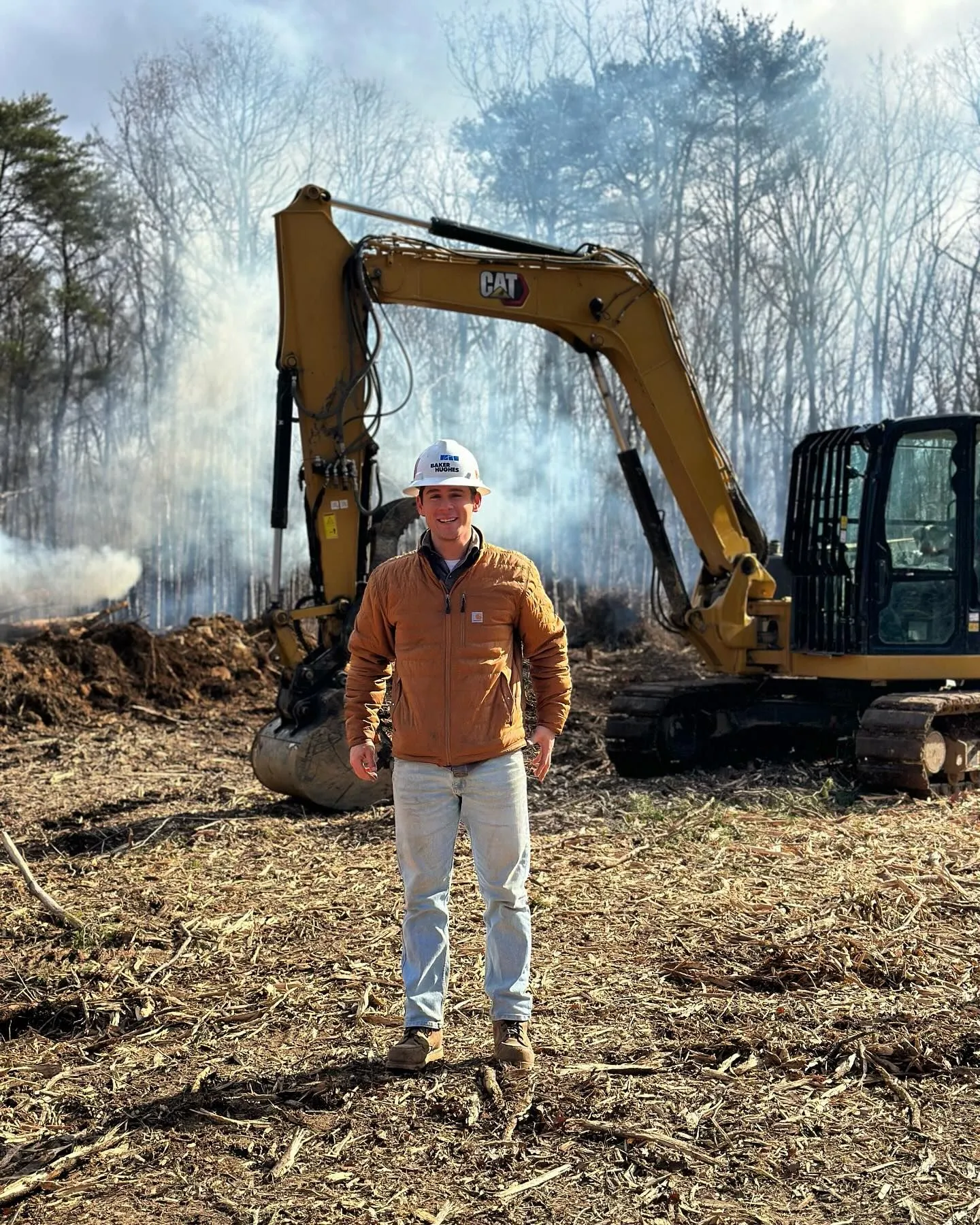 Man in hard hat stands before a CAT excavator in a clearing, with brush burning in the background.