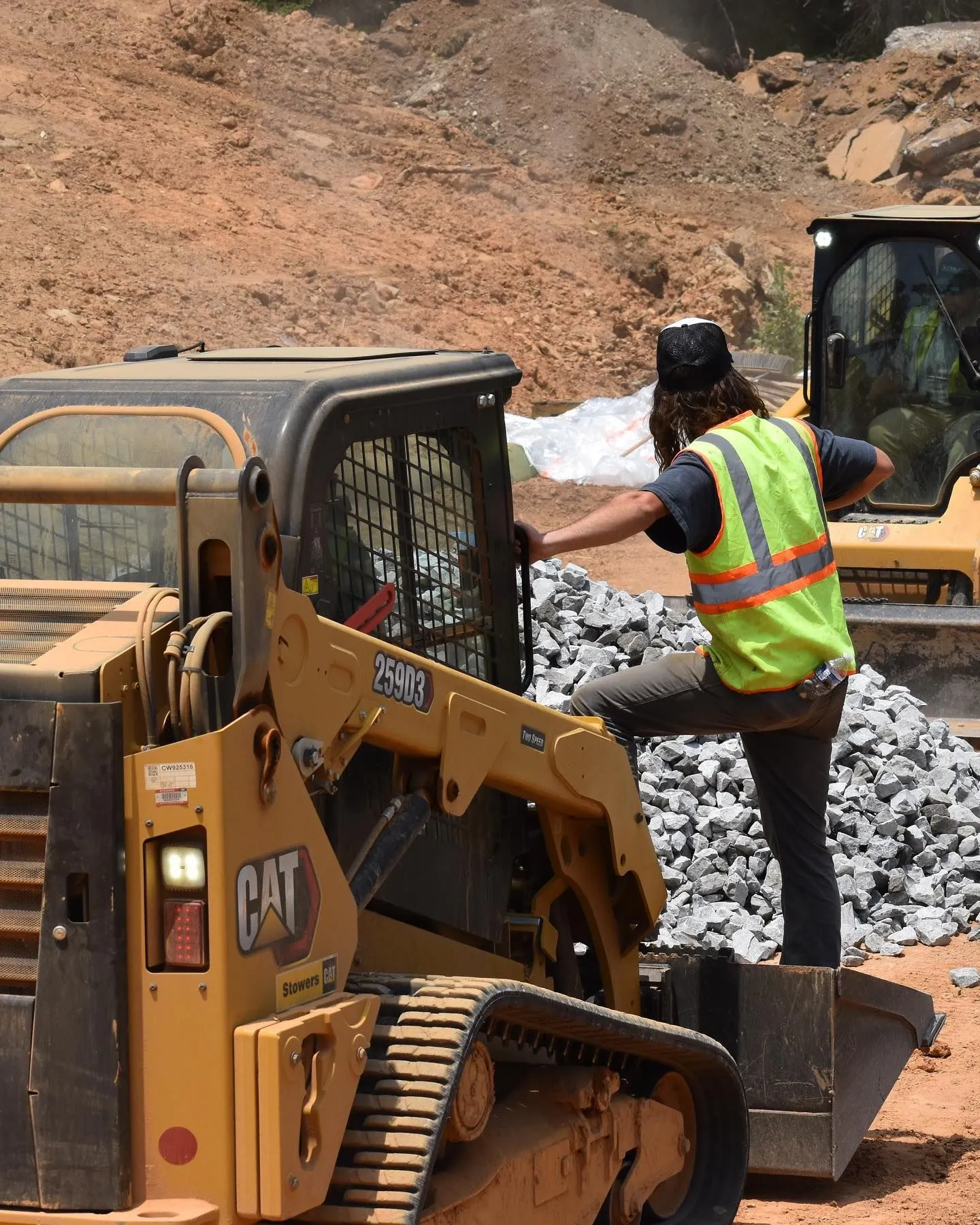 Operator climbing into a Cat 259D3 track loader beside piles of crushed gravel