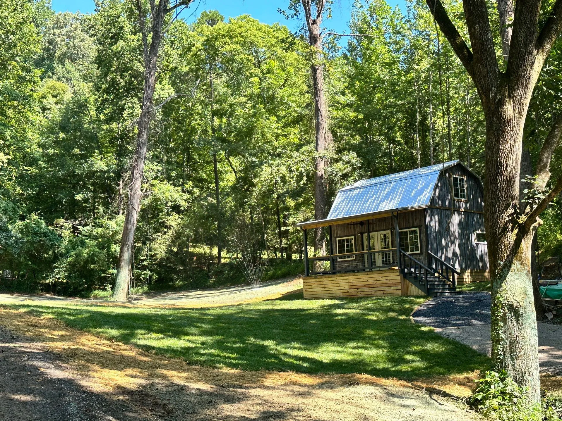 Small, dark cabin with porch in a wooded area; green grass and trees surround it.
