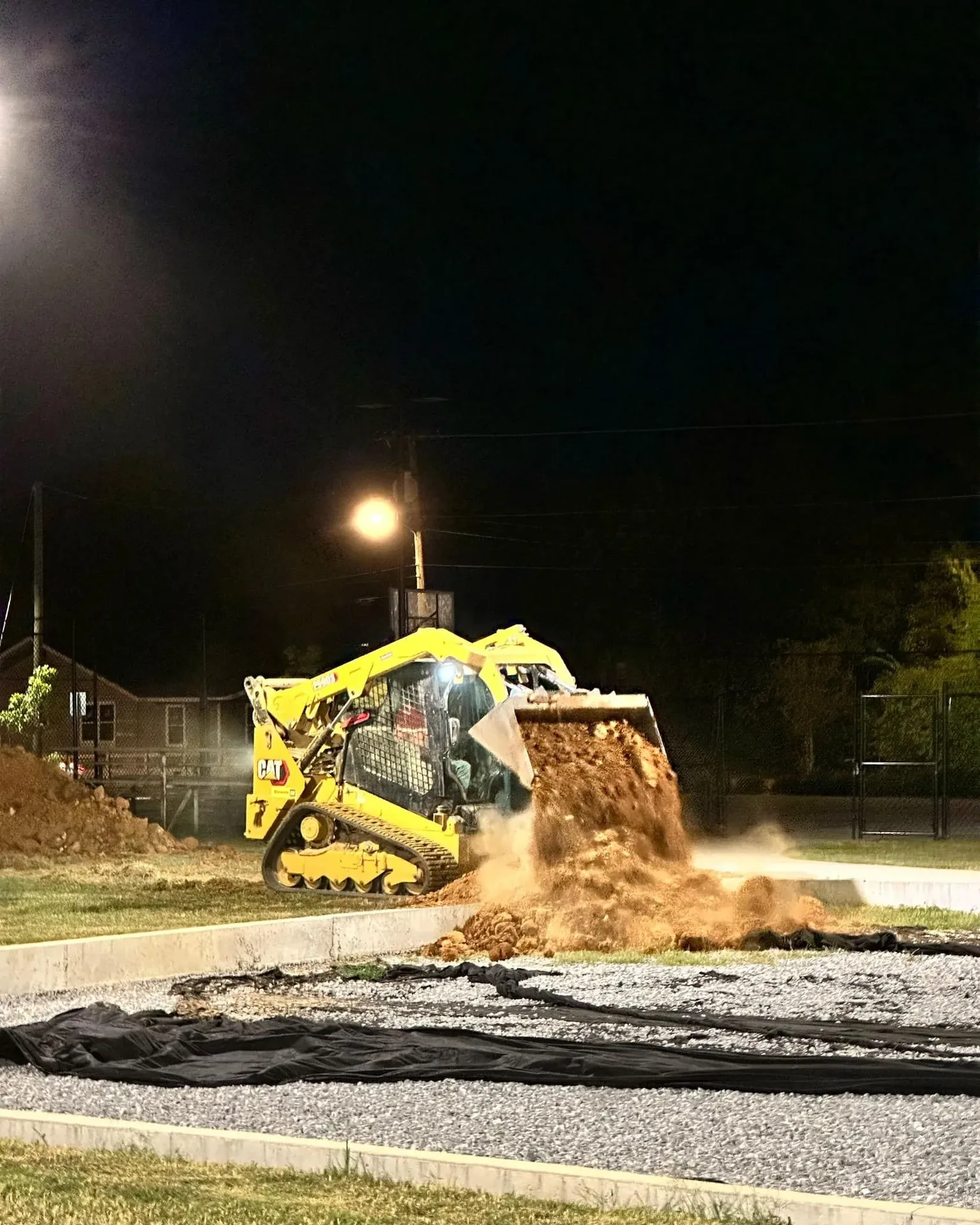Yellow skid steer dumping soil onto a dark area at night; a single streetlight illuminates the scene.