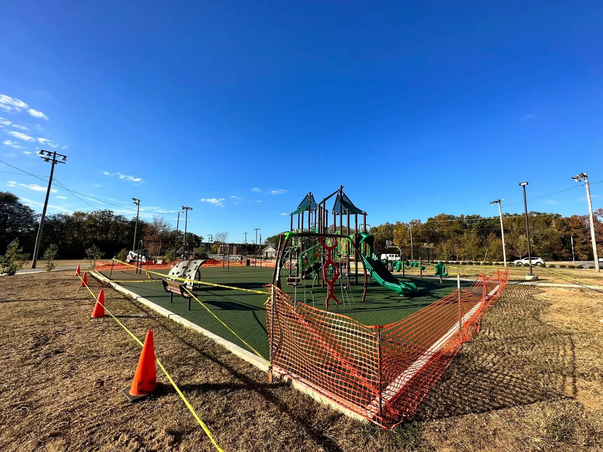 Playground under construction, orange safety fencing and cones, blue sky, and green play equipment.