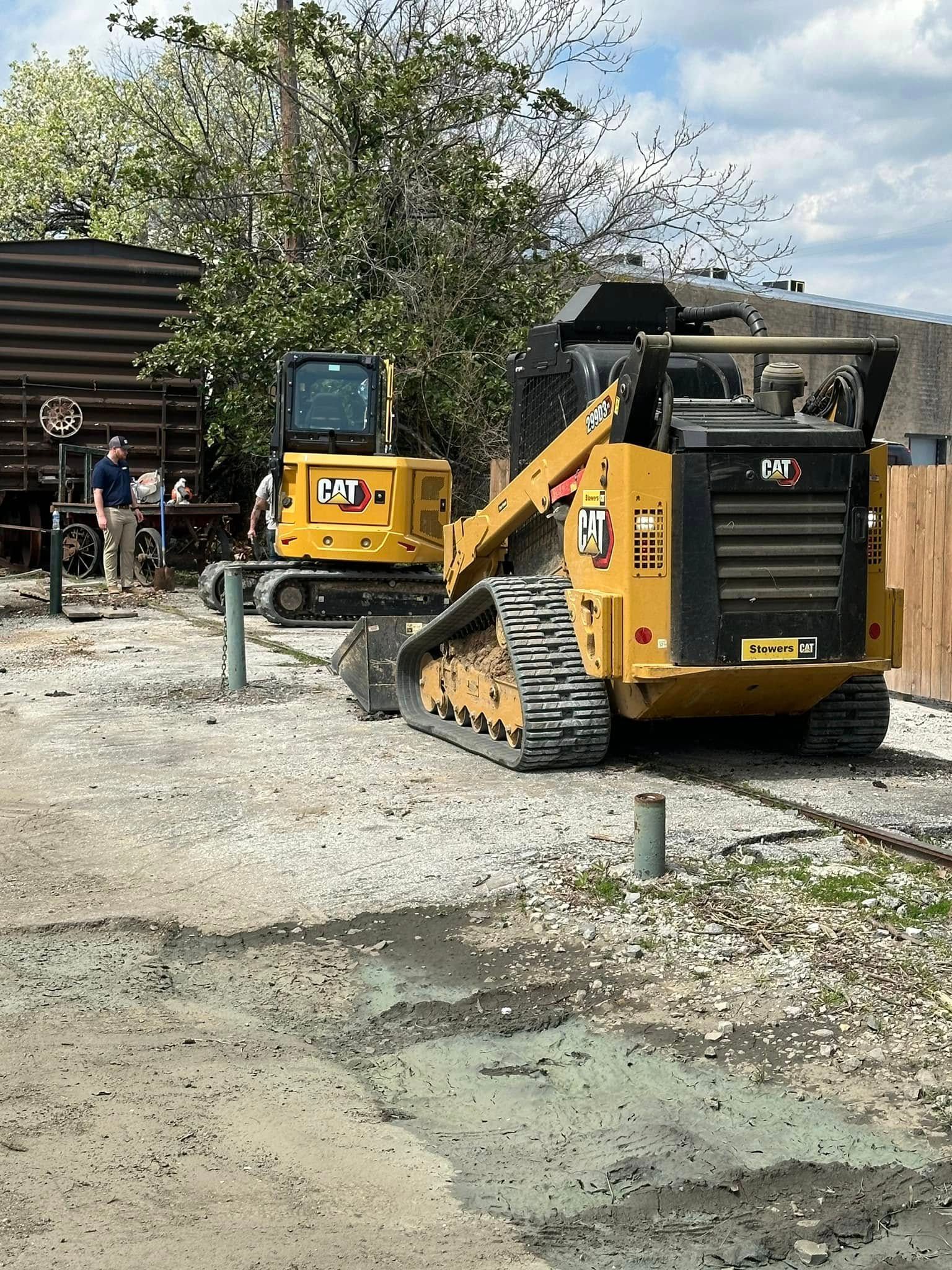 Two yellow construction vehicles on a gravel lot; a worker stands nearby.
