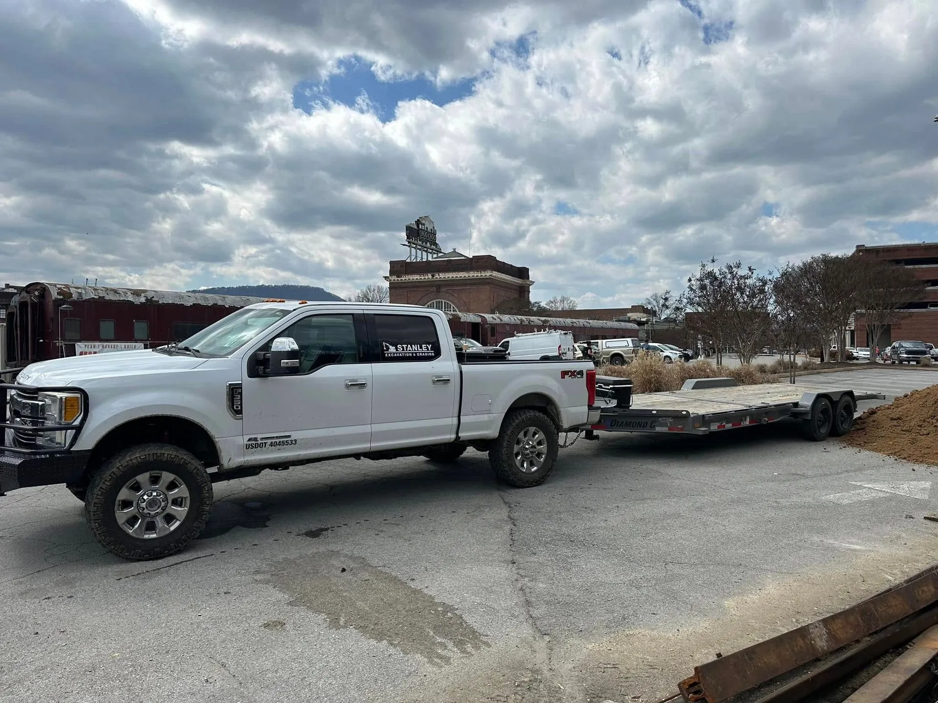 White pickup truck pulling an empty flatbed trailer in a parking lot, with a cloudy sky and brick buildings in the background.