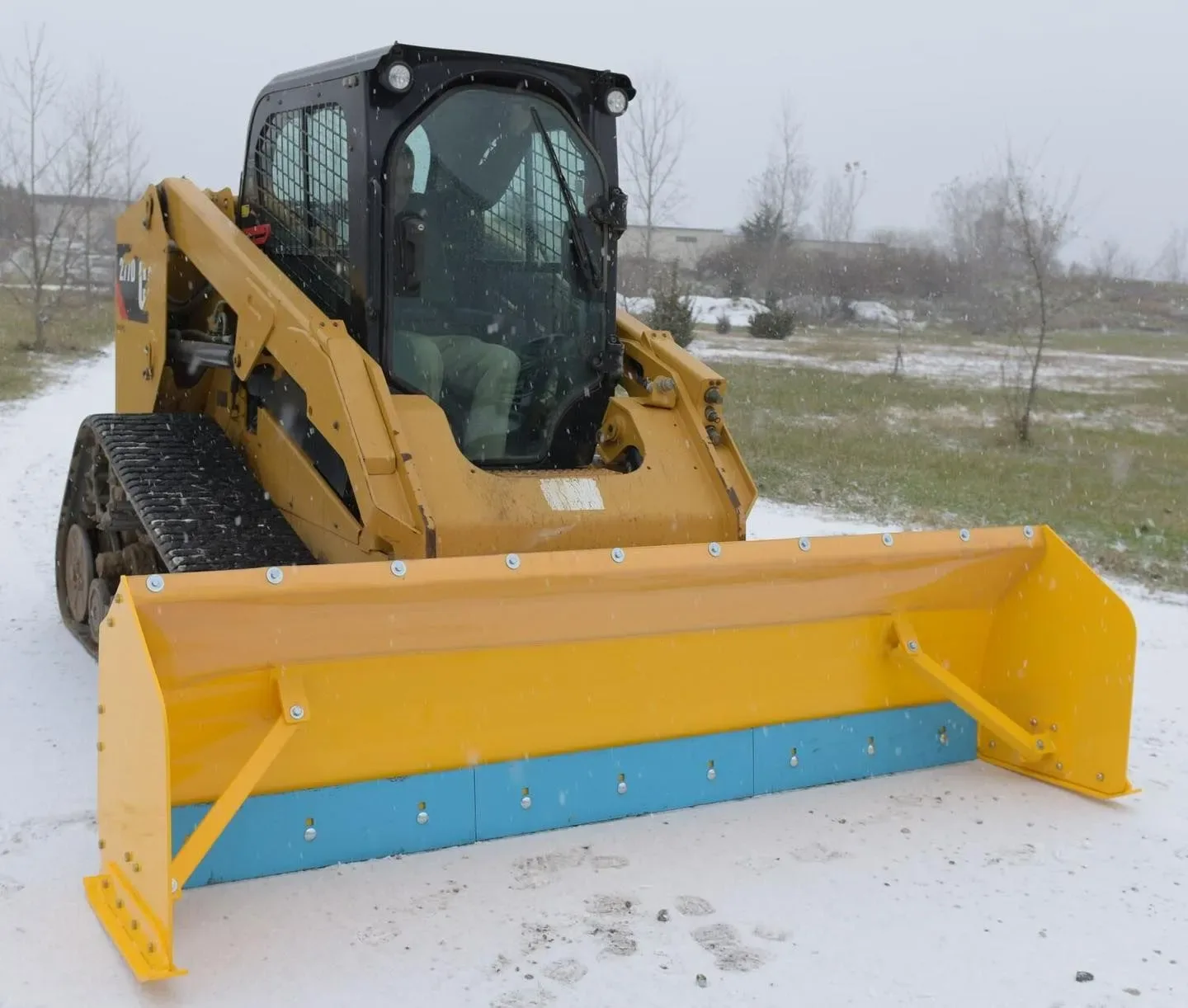 Yellow skid-steer loader with snowplow, clearing a snowy path outdoors.