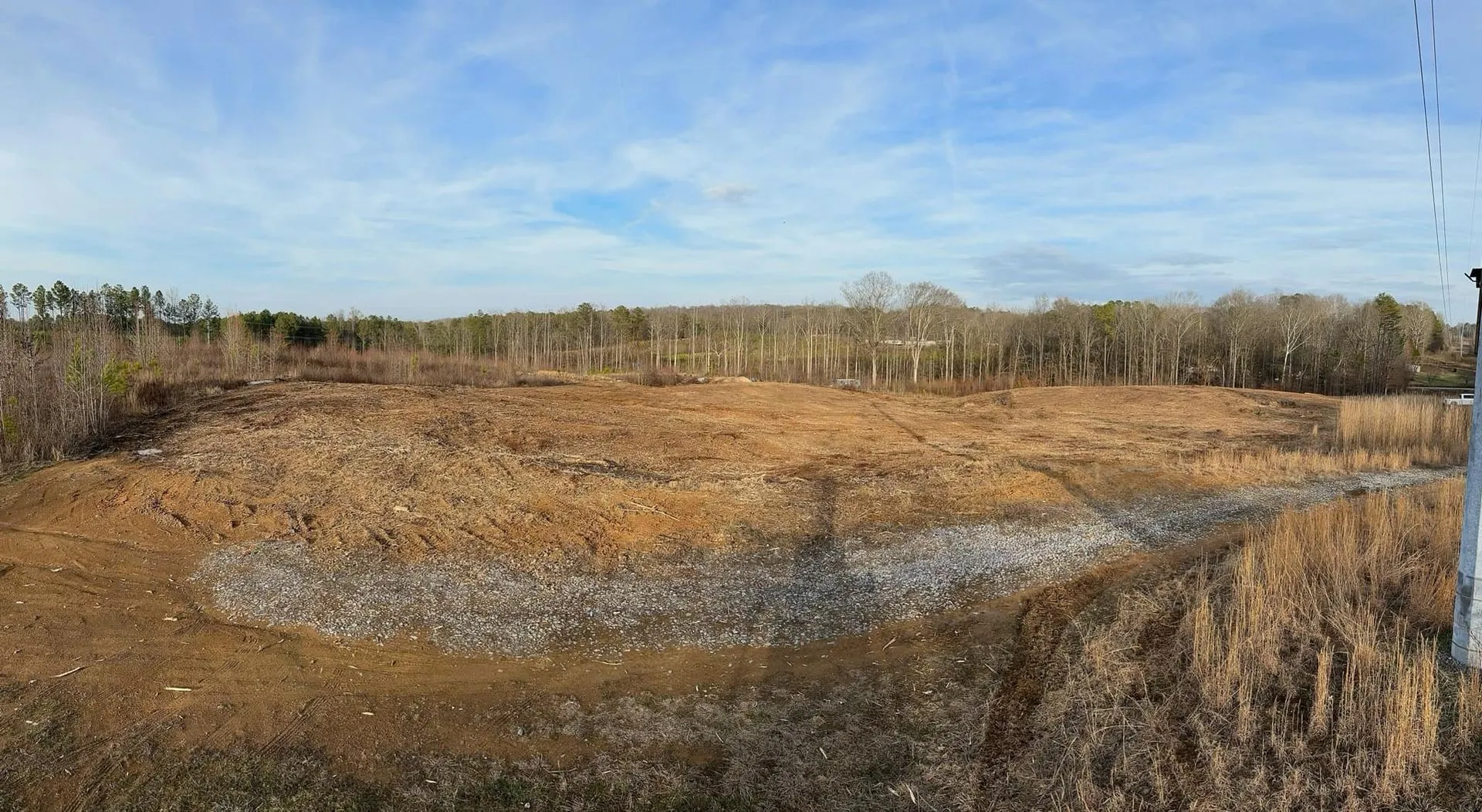 Barren, brown field with small rocks and dry brush, edged by a line of trees against a blue sky.