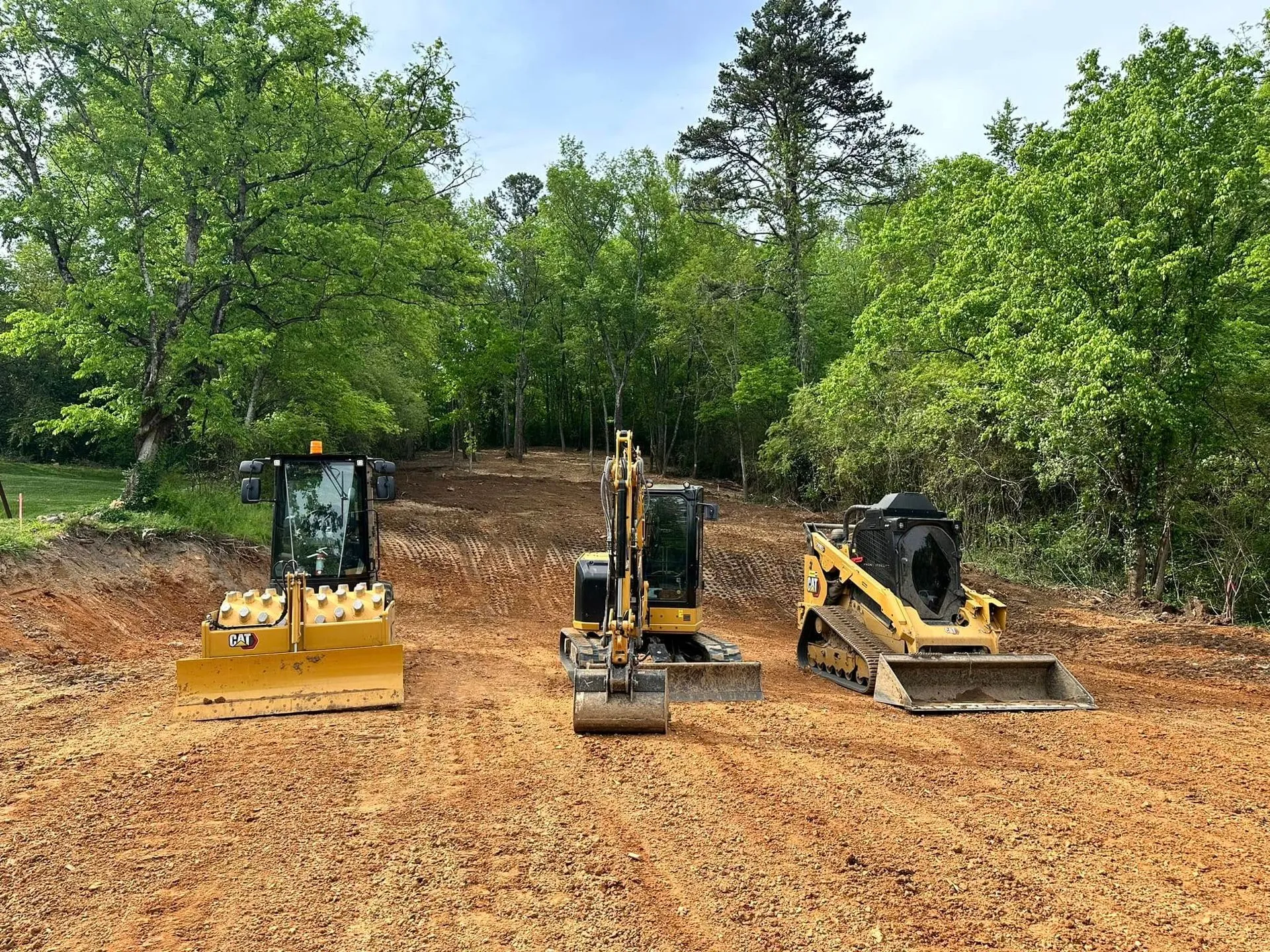 Three pieces of construction equipment on dirt with trees in the background.