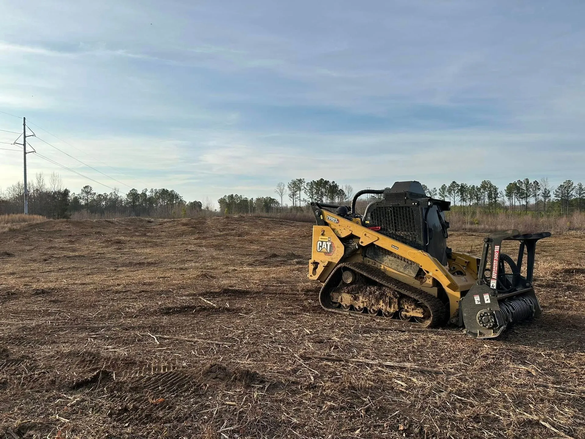 Yellow skid steer clearing a field, blue sky, trees in the background.