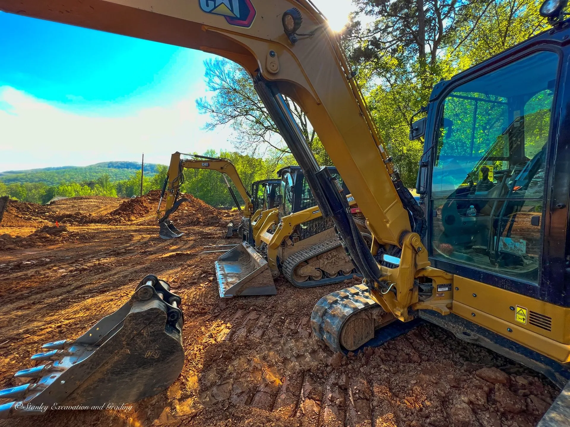 Yellow CAT excavators on muddy ground; construction site with blue sky and trees in the background.