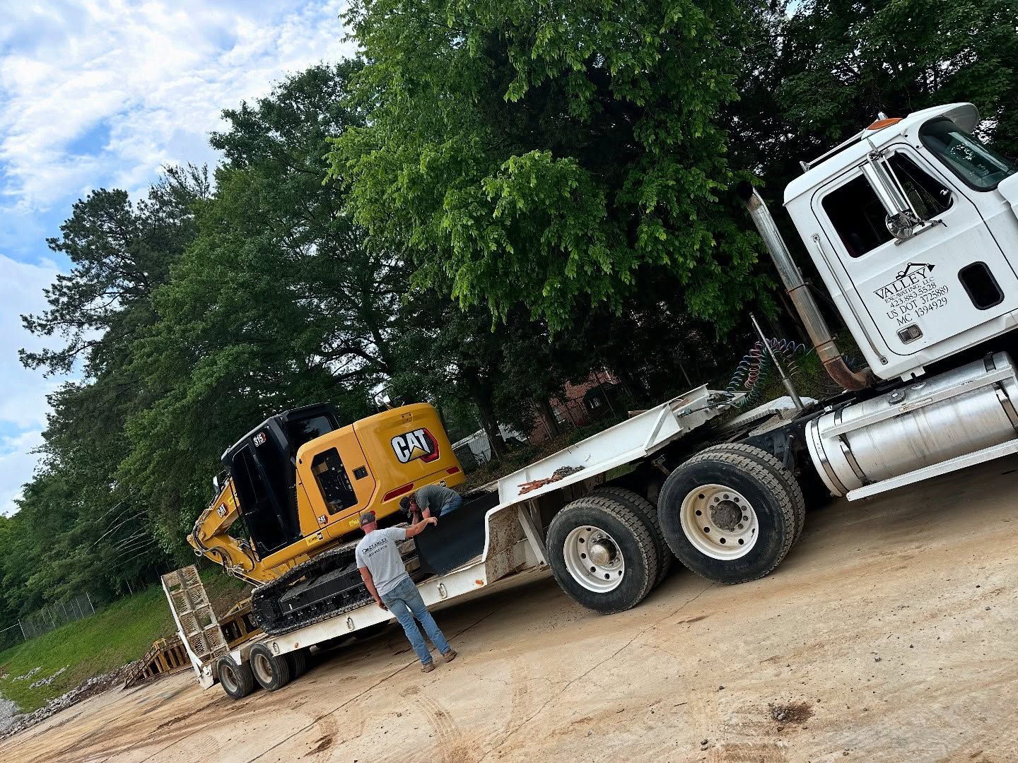 Yellow CAT excavator on a flatbed trailer being loaded by a person; a semi-truck is in the background.