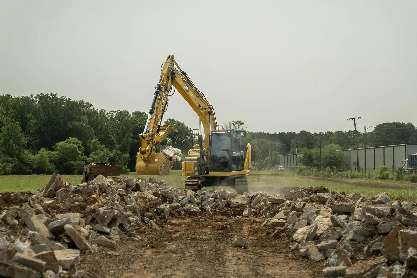 Yellow excavator demolishing concrete rubble in a field, trees in the background.