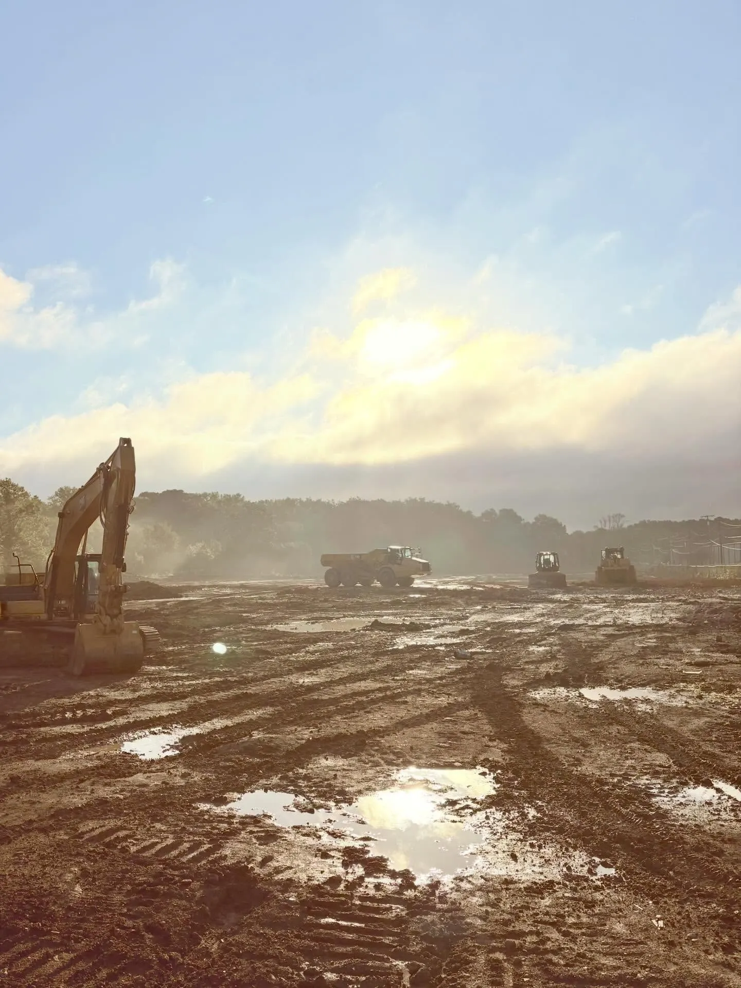 Construction site; excavator and trucks on muddy ground under a hazy sky.
