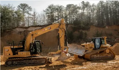 Construction site with dirt, gravel, and trees under a blue sky, an excavator works in the distance.