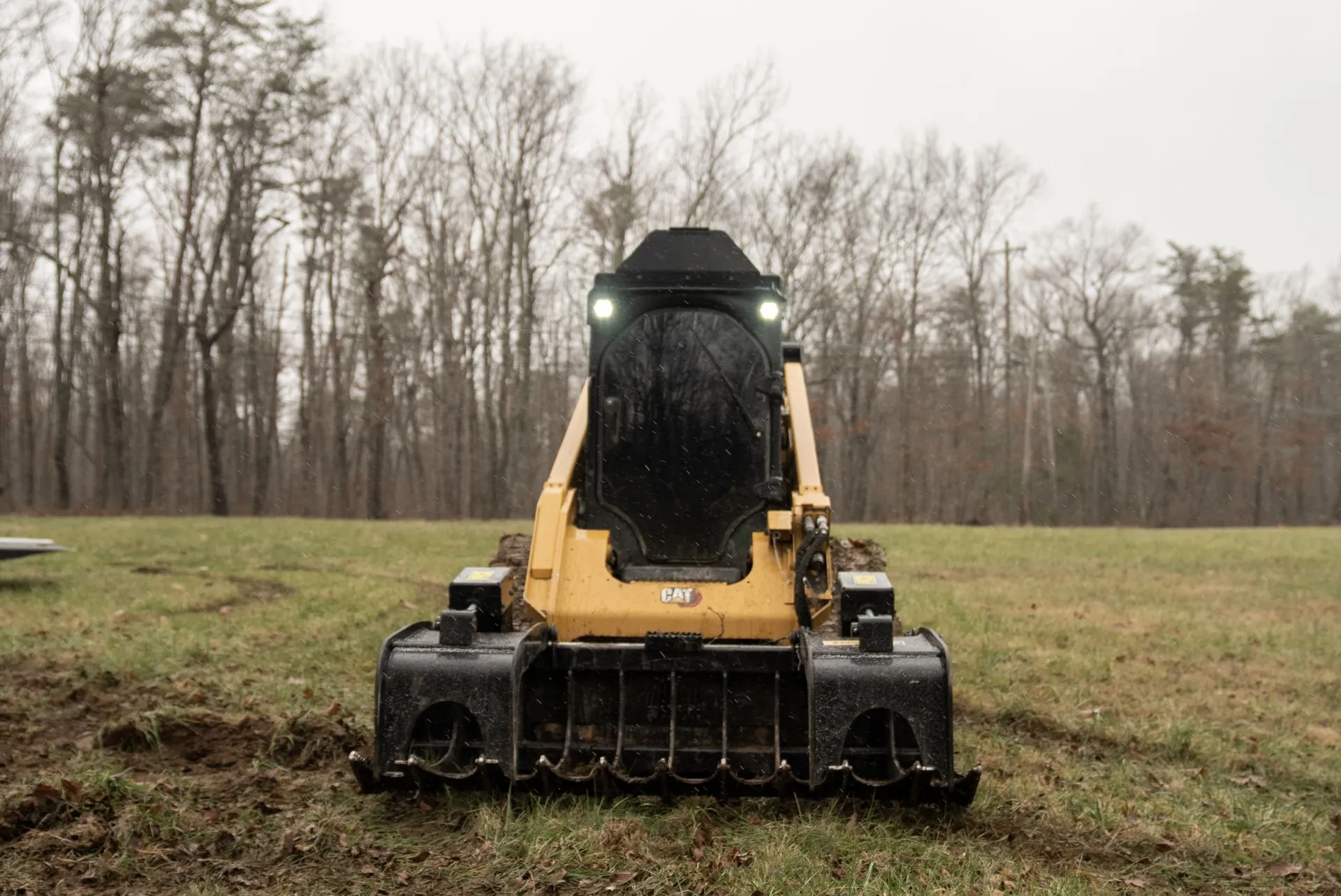 Yellow skid steer with front attachment on muddy field, trees in the background, possibly rainy day.
