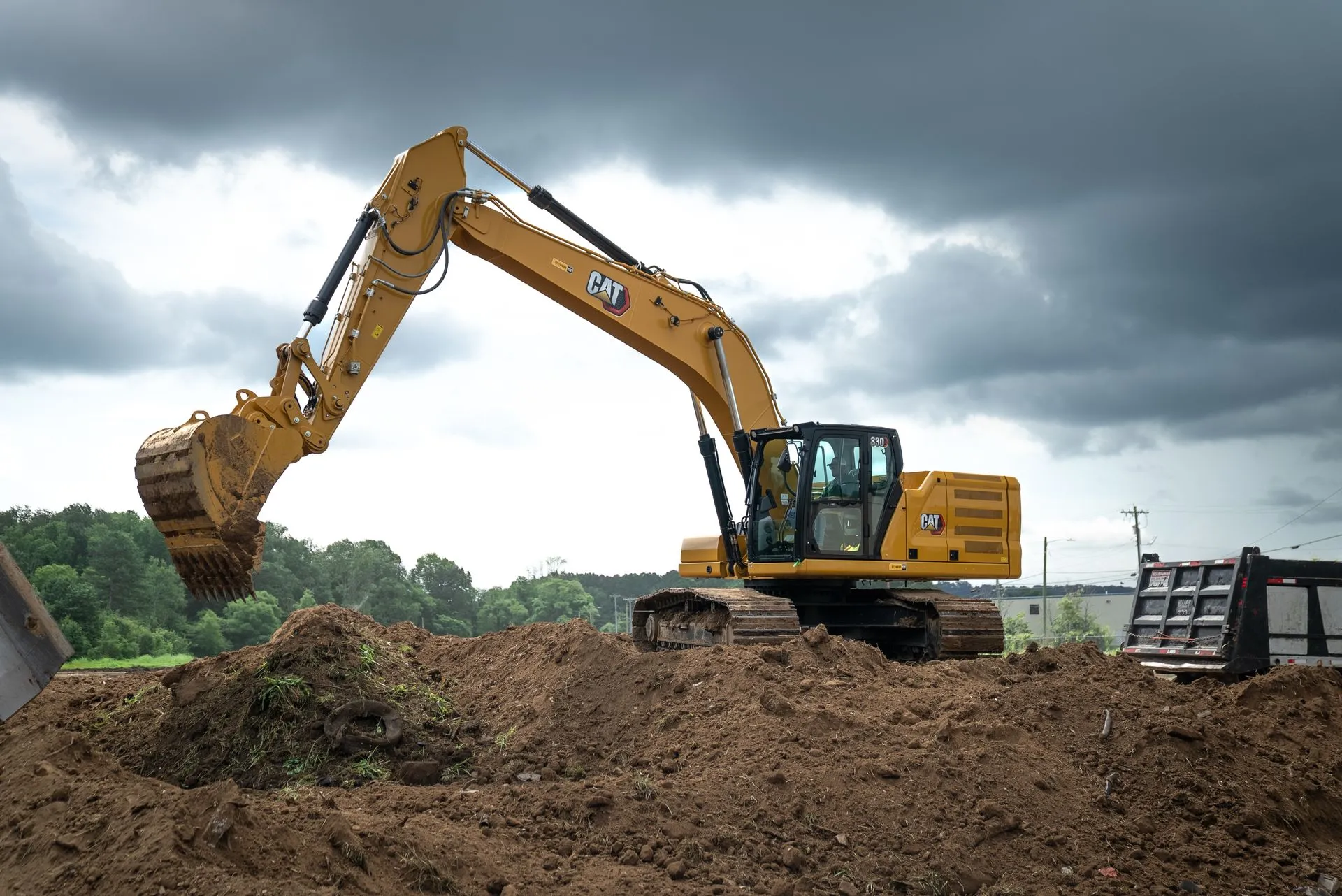Yellow Caterpillar excavator scoops earth from a dirt pile under a cloudy sky.