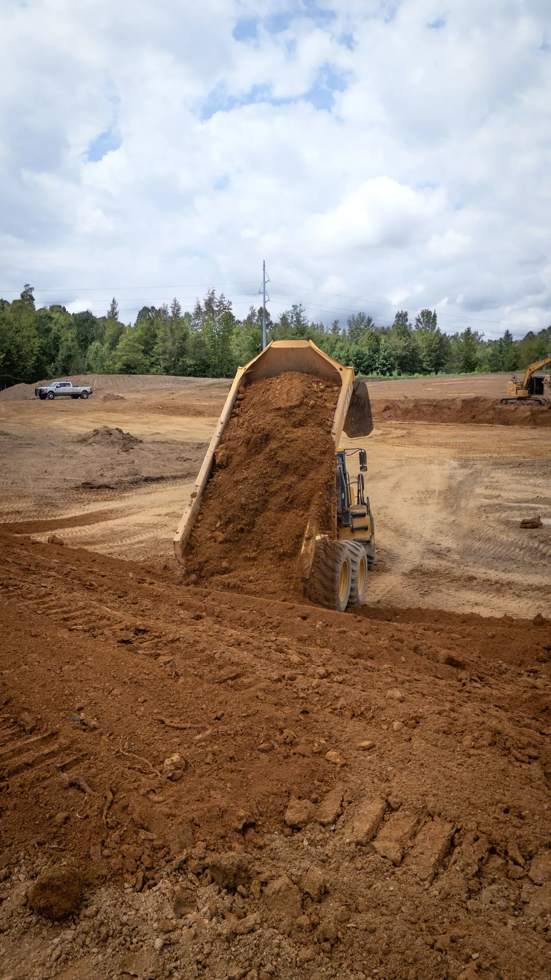 Dump truck dumping brown soil onto a construction site under a cloudy sky.