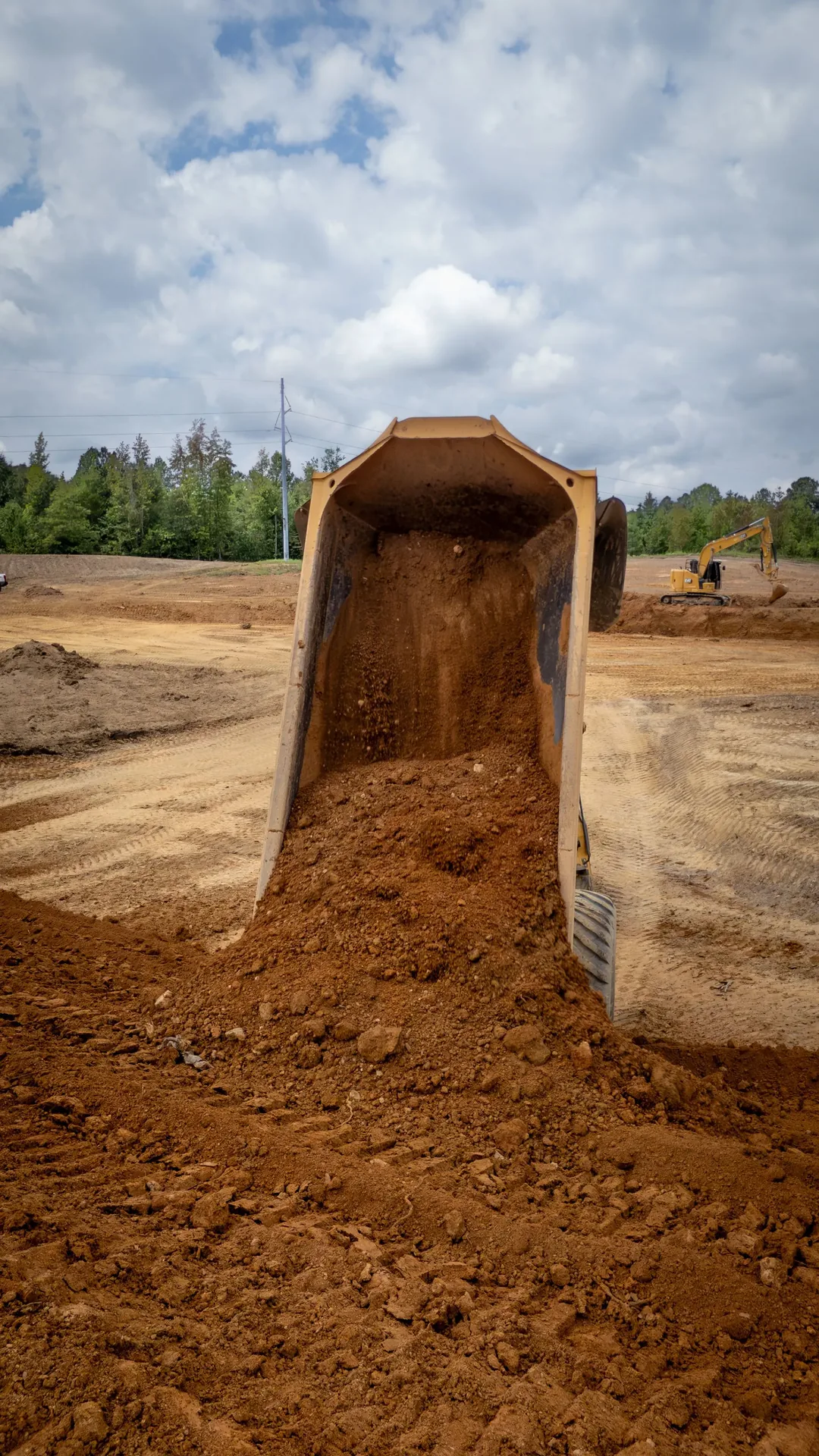 Dump truck bed tilted, dumping brown soil onto dirt ground. Blue sky with clouds.