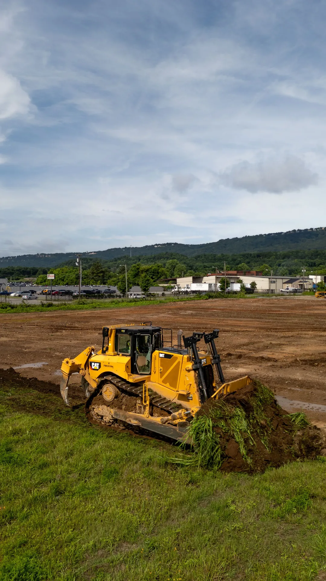Yellow bulldozer pushing dirt and vegetation, with a landscape background under a cloudy sky.