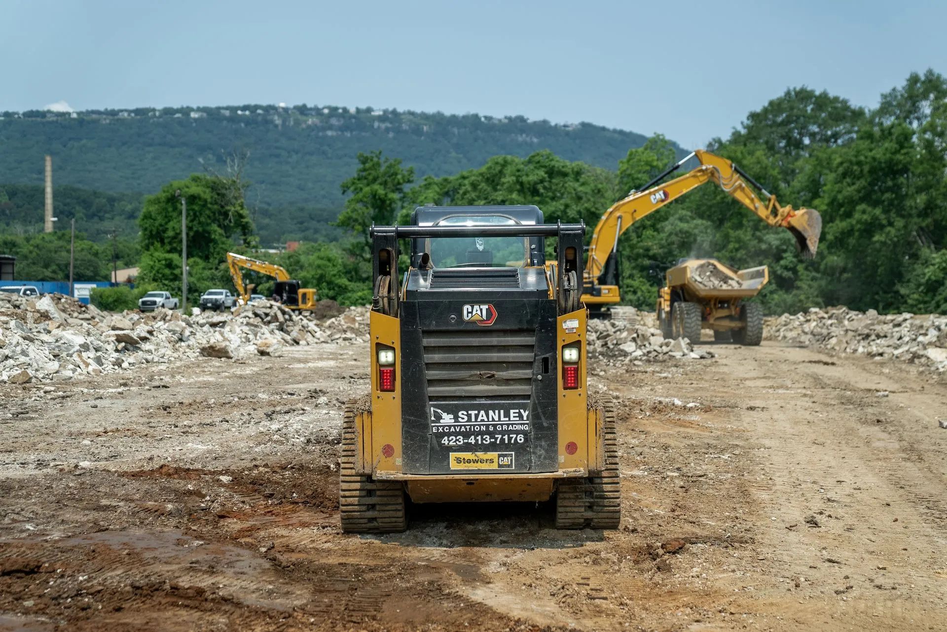 Rear view of a yellow Caterpillar skid steer at a construction site with other machinery; a hill in the background.