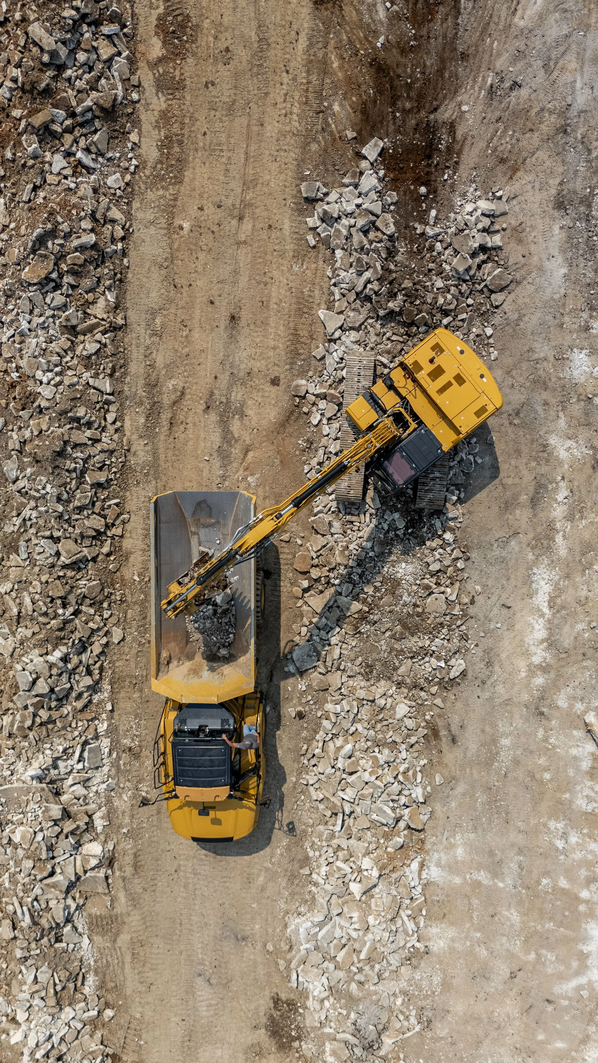 Overhead view: Yellow excavator loading debris into a dump truck at a waste disposal site.
