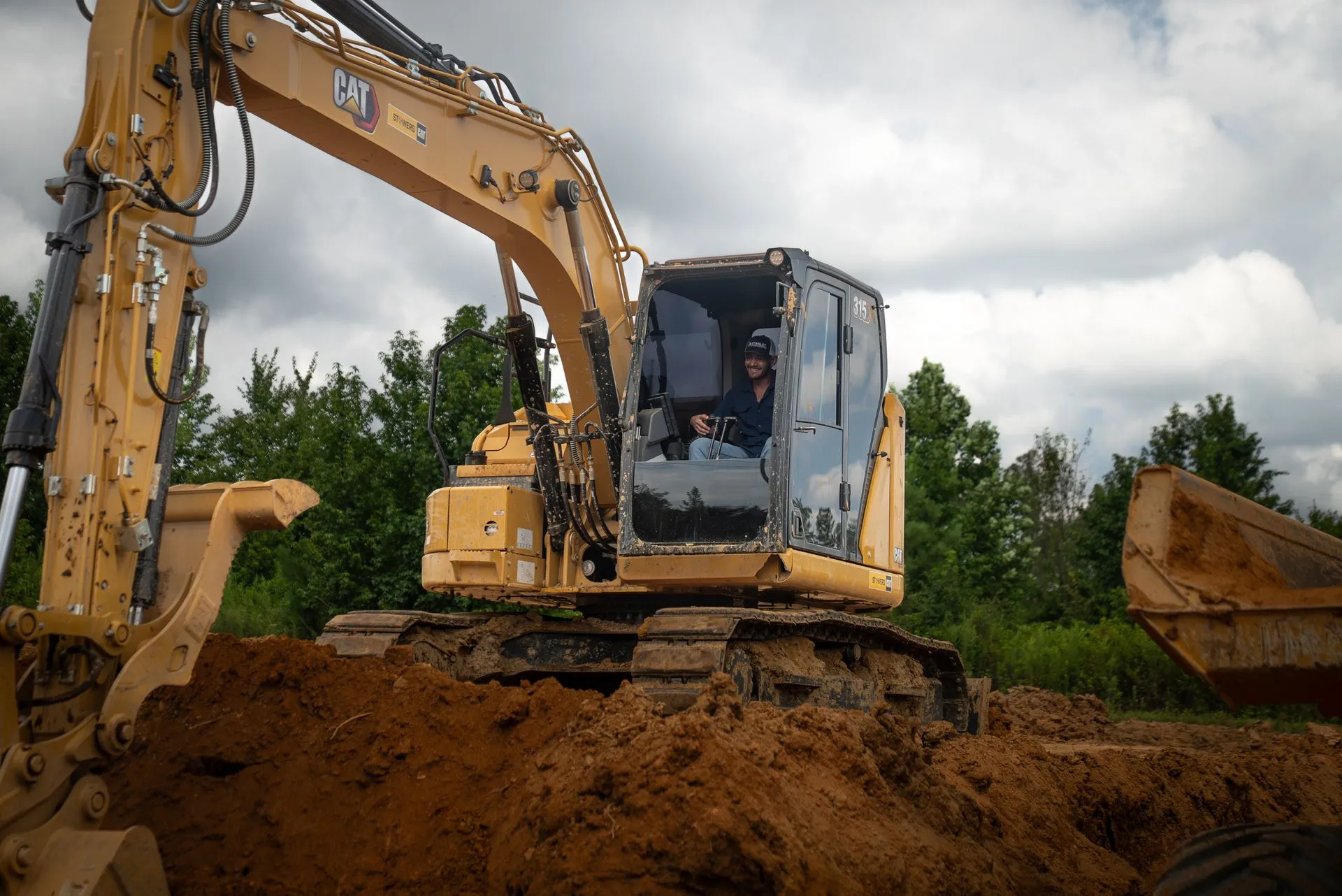 Yellow excavator on a dirt pile, operator visible in the cab. Cloudy sky, trees in the background.