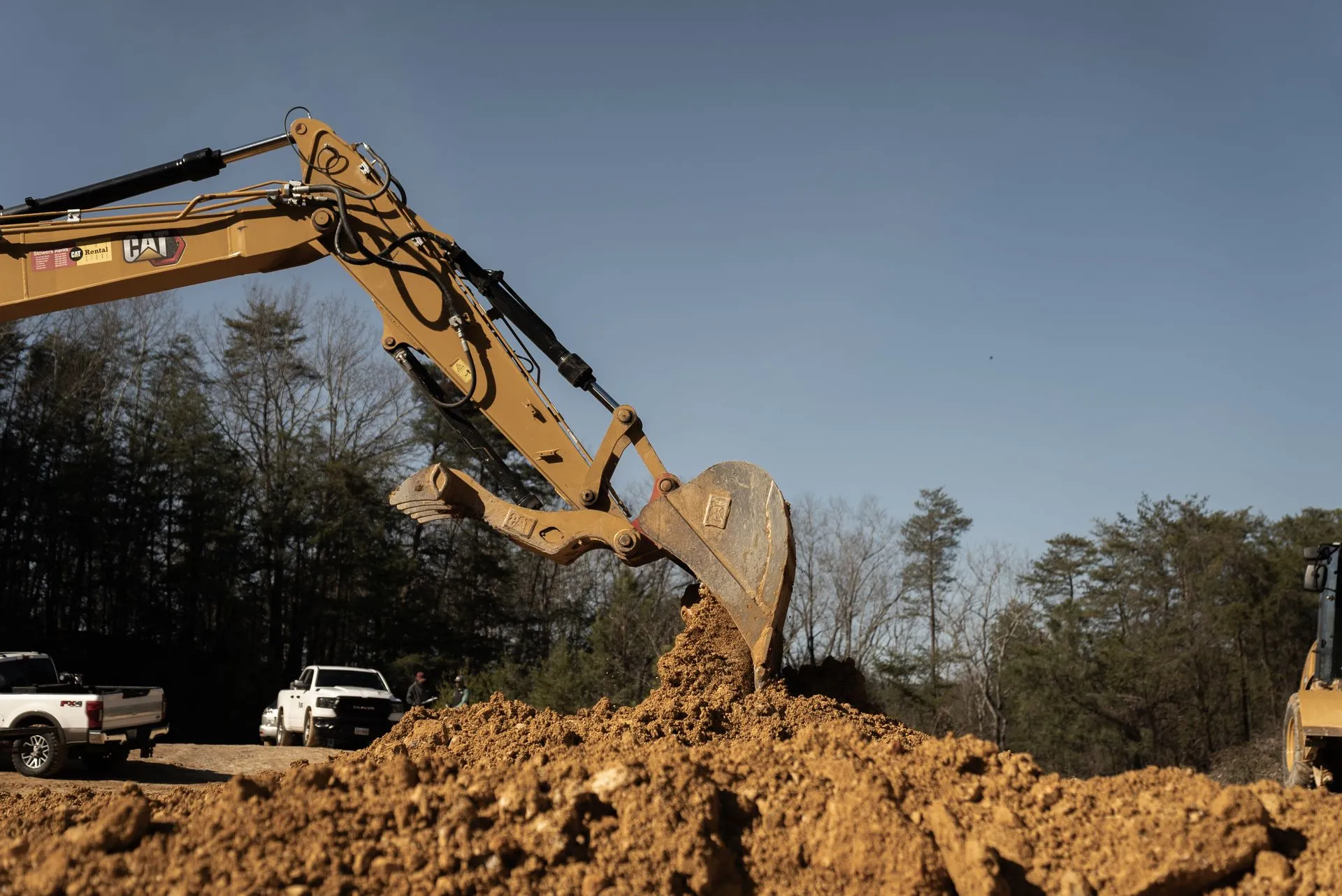 Excavator digging dirt at a construction site with white trucks and trees in the background under a blue sky.